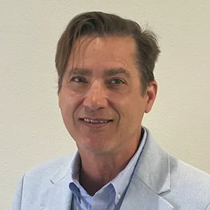 Portrait of a smiling man wearing a light blue blazer and button-down shirt, standing against a neutral light wall.