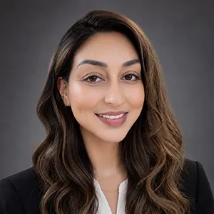 Professional headshot of a smiling woman with long dark hair, wearing a black blazer and white blouse, against a neutral gray background.