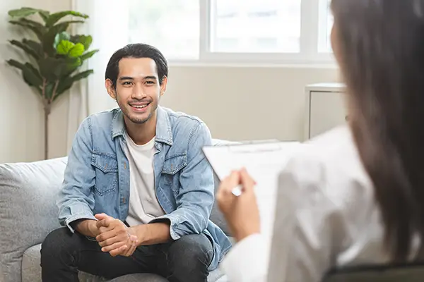 Man in a denim shirt sits on a sofa, smiling, during a counseling session with a therapist taking notes in the foreground.