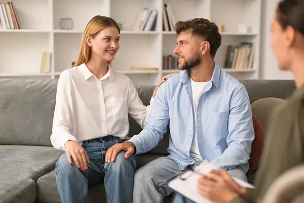 A couple sits on a sofa in a counseling session, the woman smiling and resting a hand on the man's shoulder while a therapist takes notes nearby
